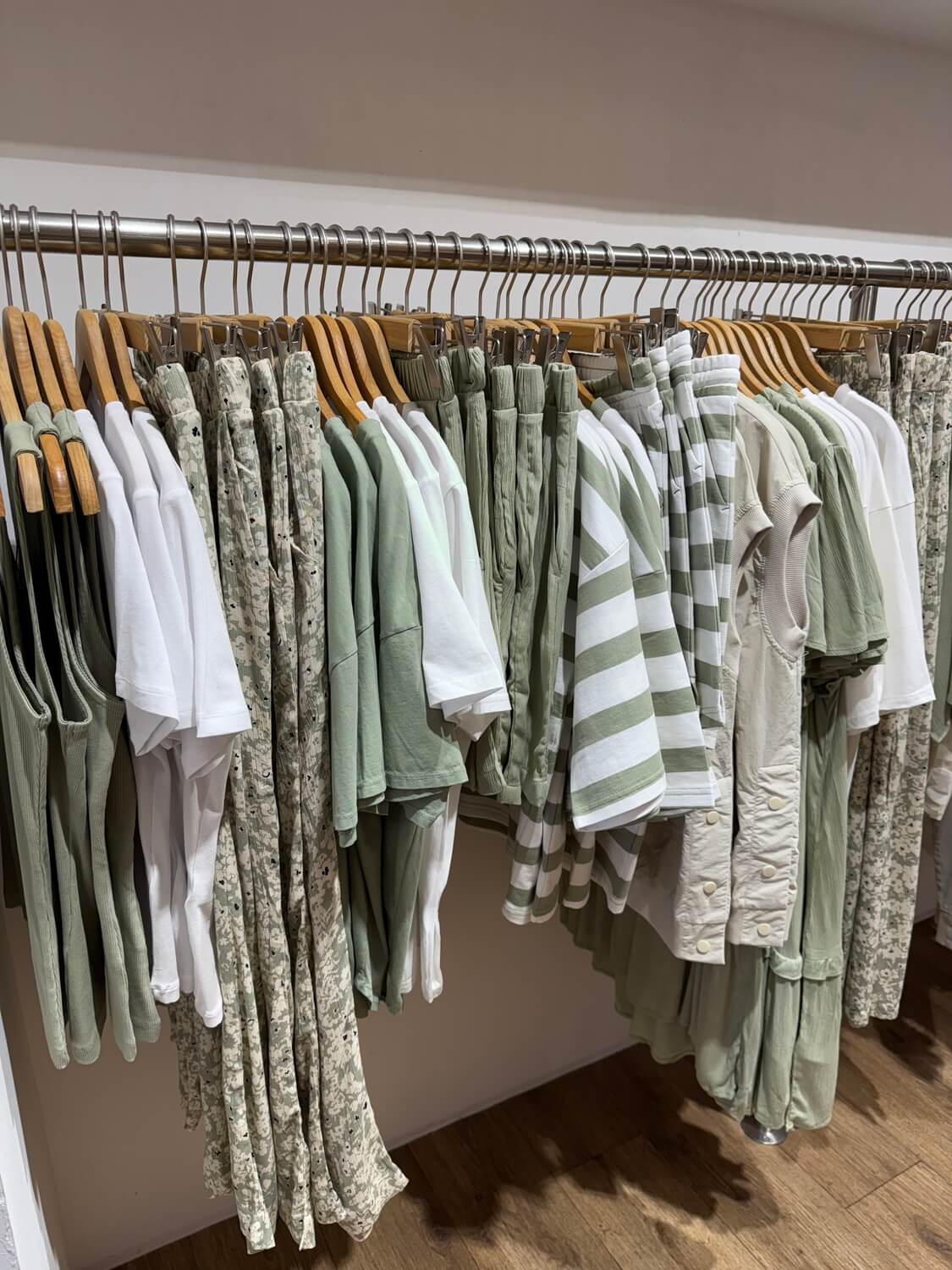 Kleiderstange mit verschiedenen Kleidungsstücken in Weiß, Grün und gemustert, darunter T-Shirts, Hemden und Hosen auf Holzkleiderbügeln.